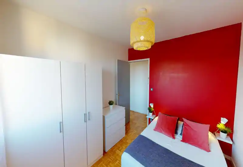 Bright, stylish bedroom with a bold red accent wall, a double bed dressed with soft pillows and a cozy throw, plus white storage furniture and warm lighting — ideal for a comfortable stay.