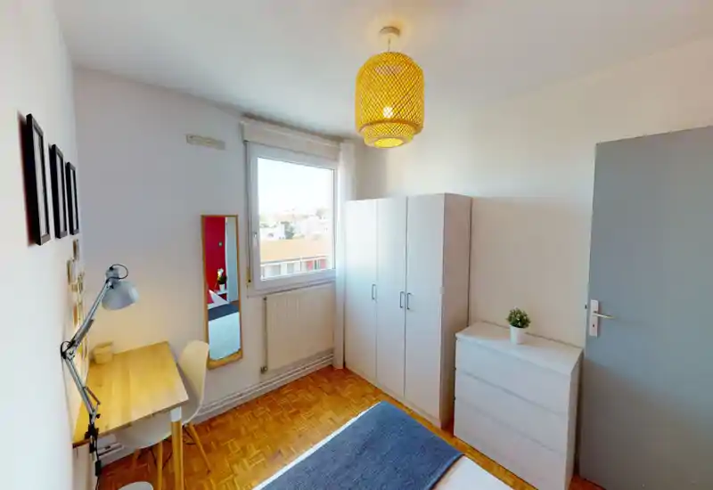 Bright, neatly staged bedroom with a visible double bed, a study desk by the wall and ample storage (wardrobe and chest of drawers). Large window provides natural light and the warm woven ceiling lamp gives a cozy atmosphere.