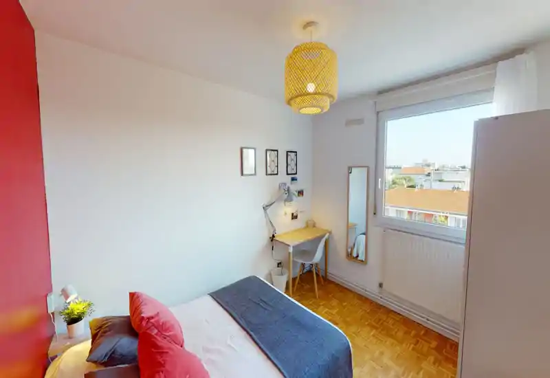 Bright, neatly staged bedroom with a double bed dressed in layered textiles, a small desk by the window and a warm woven ceiling lamp. Large window offers city views and natural light, ideal for a comfortable stay.