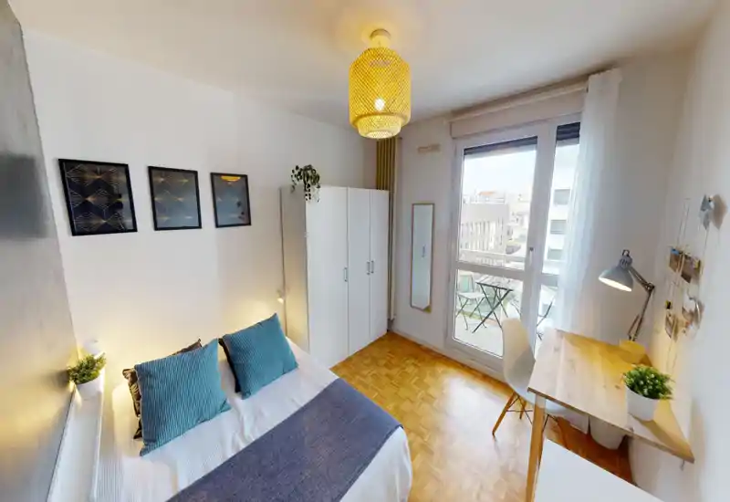 Bright, well-styled bedroom with a double bed dressed in white linens and blue accents, a wardrobe, desk by the window and a small balcony visible through large glass doors.