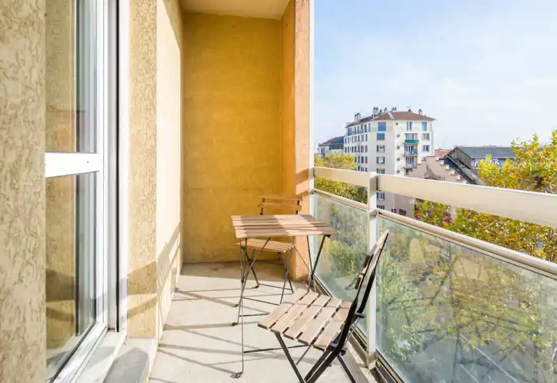 Sunny balcony with a small wooden bistro table and two foldable chairs, glass railing and a pleasant city view — ideal for morning coffee.