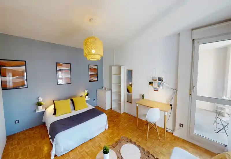 Camera da letto luminosa e ben arredata con letto ampio con biancheria bianca, cuscini gialli e lampadario intrecciato. Pavimento in parquet, scrivania vicino alla finestra e quadri alle pareti: perfetta per annunci che valorizzano comfort e luce naturale.