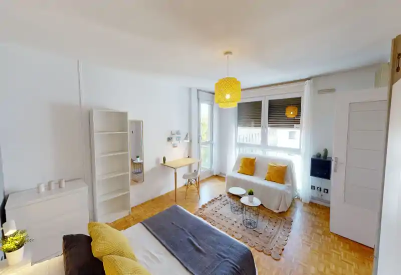 Camera da letto luminosa e moderna con letto ben sistemato in primo piano, accenti gialli coordinati, una piccola scrivania vicino alla finestra e un'area salotto accogliente — ideale per una persona o una coppia.