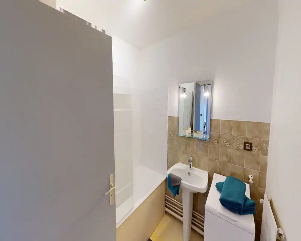 Compact bathroom with a bathtub/shower combination, pedestal sink, mirror and washing machine. Neutral tiles and folded towels create a tidy, functional space.