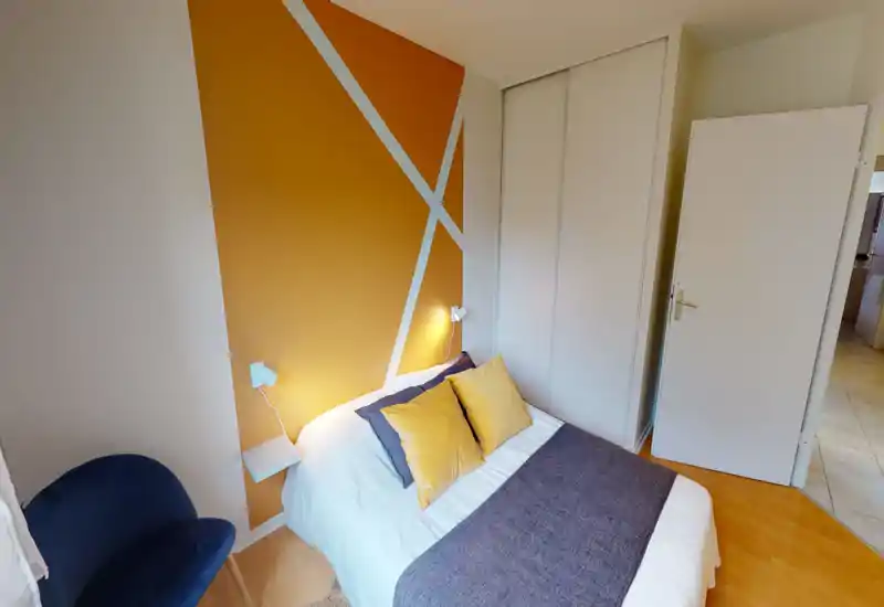 Chambre moderne et lumineuse avec un lit double, coussins décoratifs et un mur d'accent jaune à motifs géométriques ; placard intégré et fauteuil confortable.