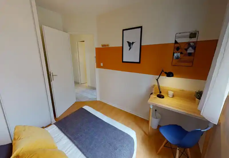 Chambre lumineuse et bien aménagée avec un lit bien fait, une bande décorative orange au mur et un petit bureau en bois avec lampe — idéale pour courts séjours ou étudiants.