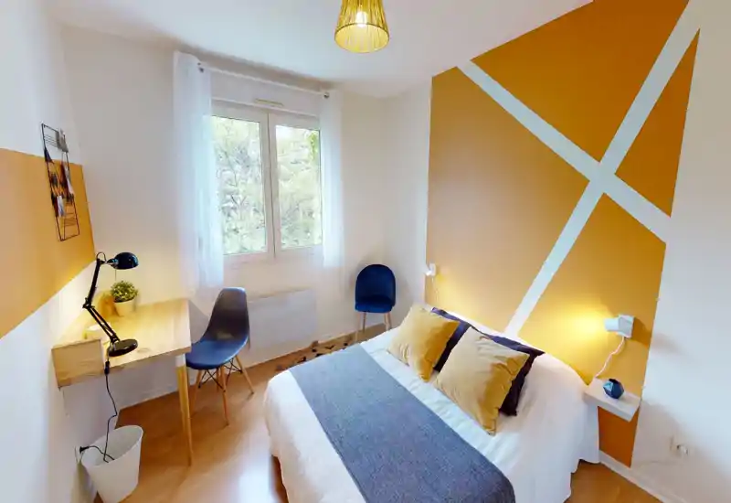 Chambre moderne et lumineuse avec un lit double confortable, des coussins décoratifs et un mur d'accent géométrique. Un espace de travail avec bureau en bois et lampe près de la fenêtre apporte fonctionnalité et convivialité.