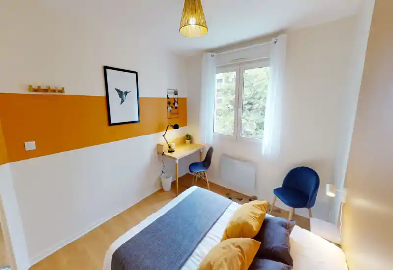 Chambre lumineuse et soignée avec un lit double confortable, des accents jaunes chaleureux et un petit bureau près de la fenêtre — idéale pour se détendre ou travailler.