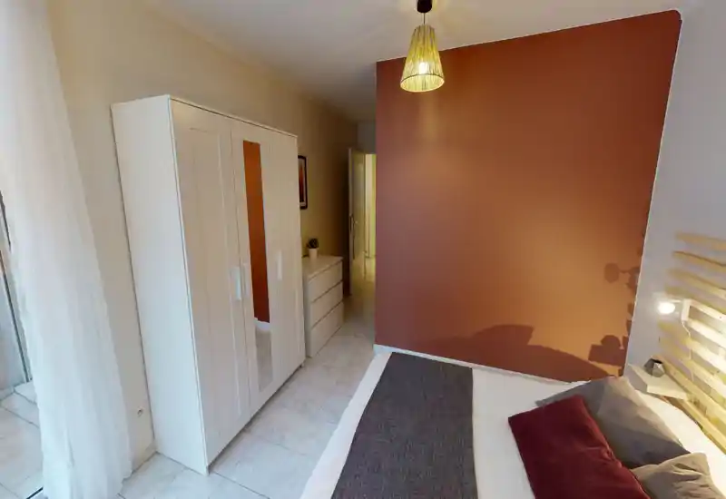 Camera da letto luminosa e ben arredata con letto ampio parzialmente visibile, parete accentuata color terracotta, lampada da comodino e armadio bianco con spazio contenitivo.