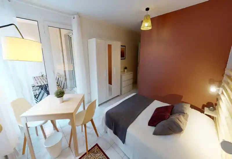 Camera da letto luminosa e ben arredata con letto matrimoniale, tessili morbidi e balcone visibile attraverso ampie porte finestre. Arredi neutri e una parete d'accento calda creano un ambiente accogliente e moderno, ideale per affitti brevi.