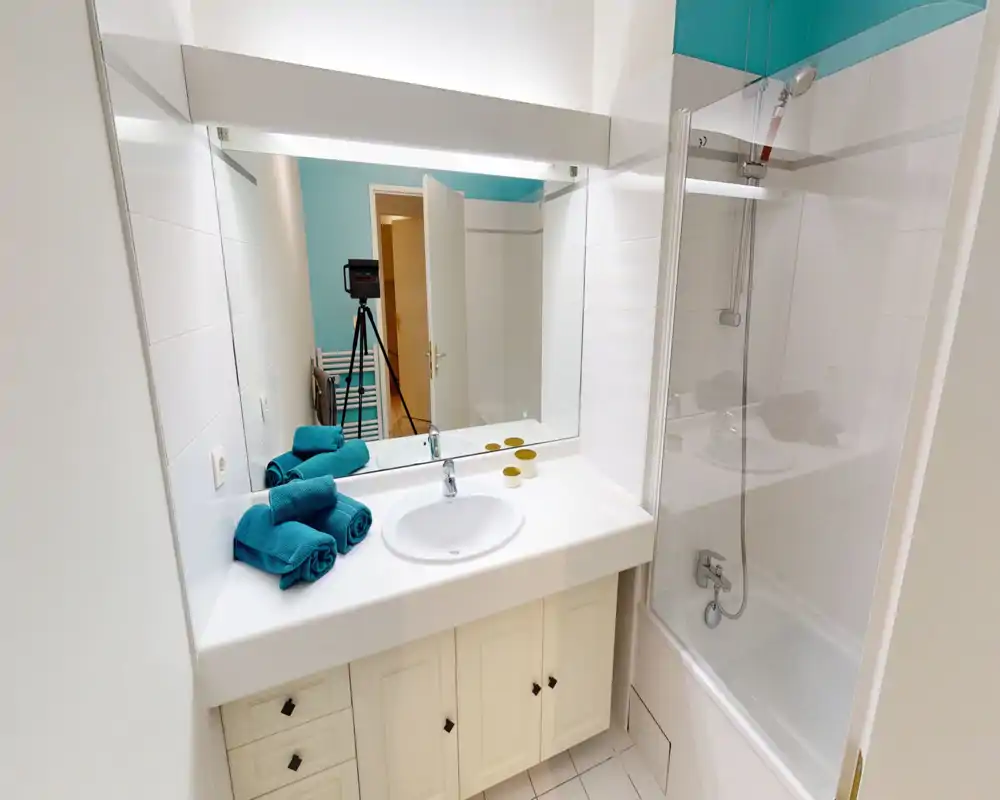 Salle de bains propre et lumineuse avec lavabo, grand miroir et baignoire avec douche. Des serviettes turquoise apportent une touche fraîche; l'espace est bien entretenu et bien éclairé.