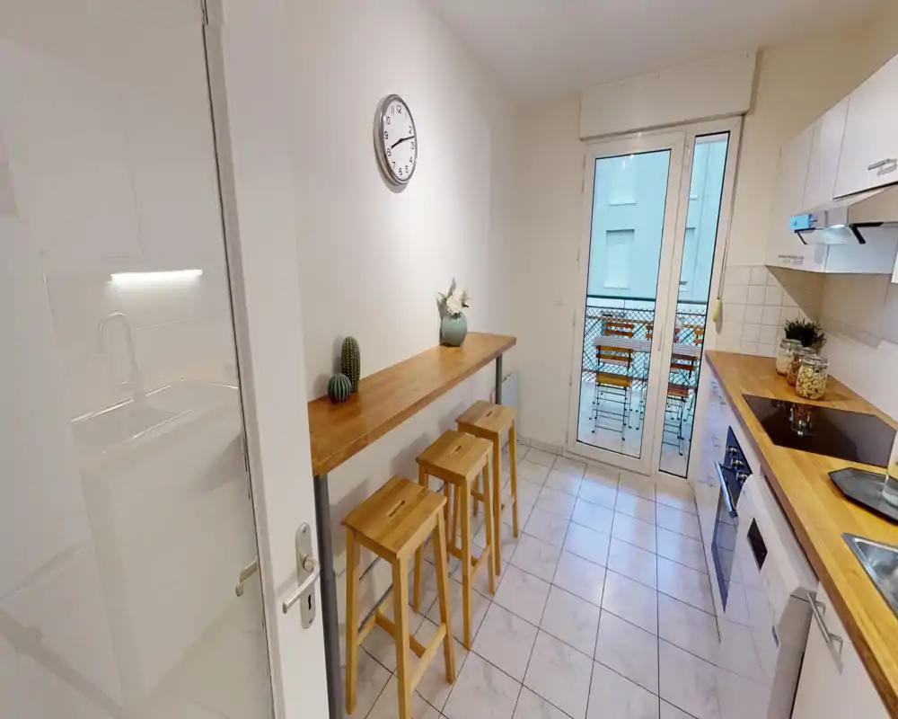 Cuisine moderne et lumineuse avec plans de travail en bois, four encastré et un petit bar avec trois tabourets; grande porte vitrée donnant sur un balcon équipé.