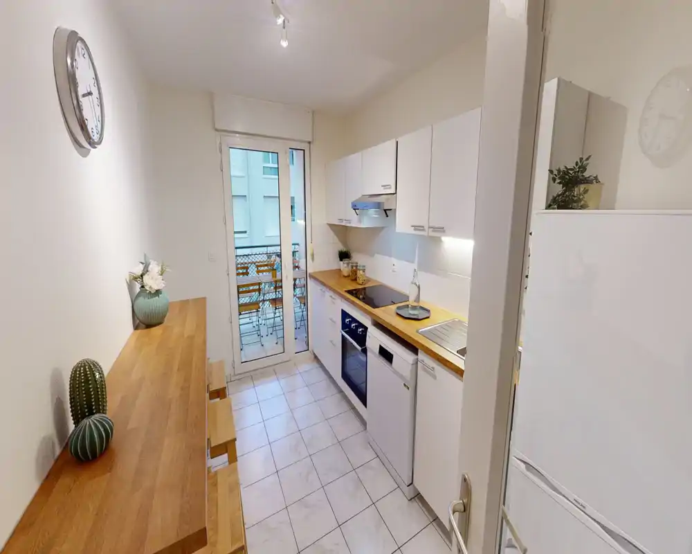 Cuisine en enfilade lumineuse et moderne avec meubles blancs, plans de travail en bois et appareils encastrés ; porte donnant sur un petit balcon.