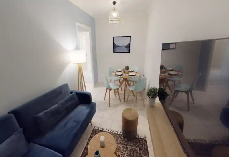 Pièce de vie lumineuse et décorée avec un canapé bleu, une lampe sur pied et une petite table à manger pour quatre. Sol carrelé, tapis décoratif et TV pour un salon moderne et confortable.