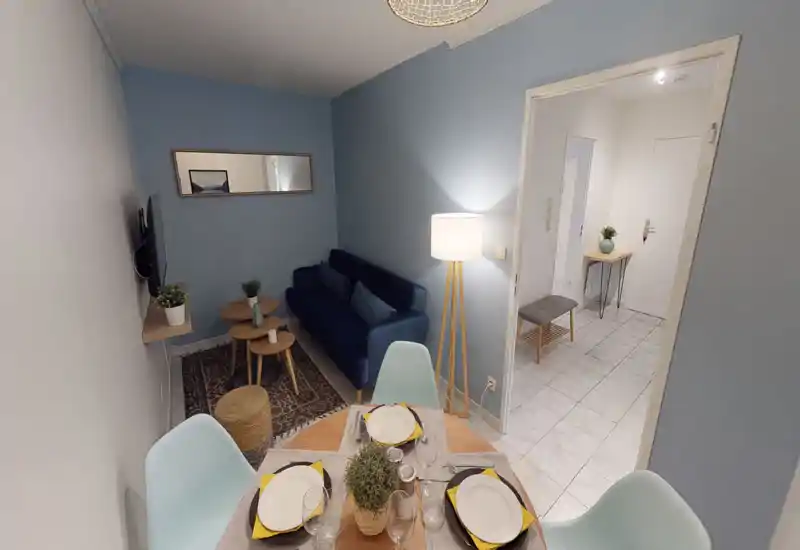 Espace salon lumineux et bien mis en scène avec un canapé bleu marine, des tables basses et un petit coin repas au premier plan ; éclairage doux et plantes décoratives pour une ambiance chaleureuse.