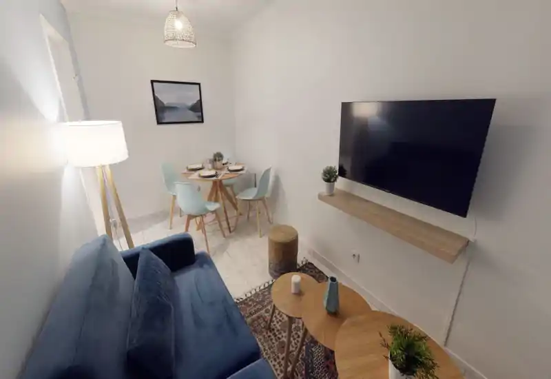 Espace salon lumineux et bien agencé avec un canapé bleu, des tables basses gigognes et une TV murale. Une petite table à manger avec des chaises pastel à l'arrière-plan crée une ambiance compacte et chaleureuse.
