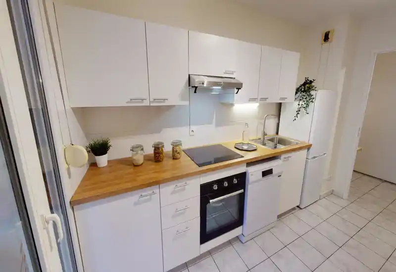 Cuisine moderne et lumineuse en enfilade avec meubles blancs, plan de travail en bois, plaque à induction et four encastré ; décoration soignée avec bocaux et petite plante.