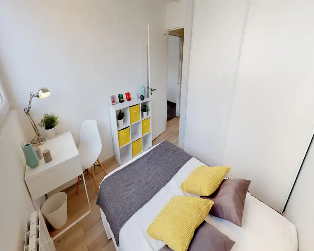 Bright, tidy bedroom with a double bed dressed in white linens, yellow accent pillows and a gray throw; a small desk and cube storage add practical workspace and organization.
