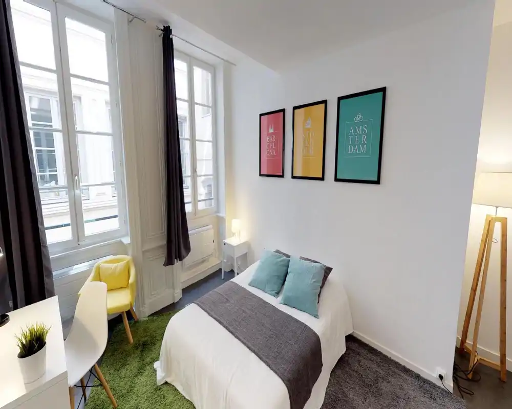 Dormitorio luminoso y bien presentado con cama doble, cojines decorativos y una manta. Grandes ventanas que aportan mucha luz natural y cuadros enmarcados que dan color a la pared blanca.