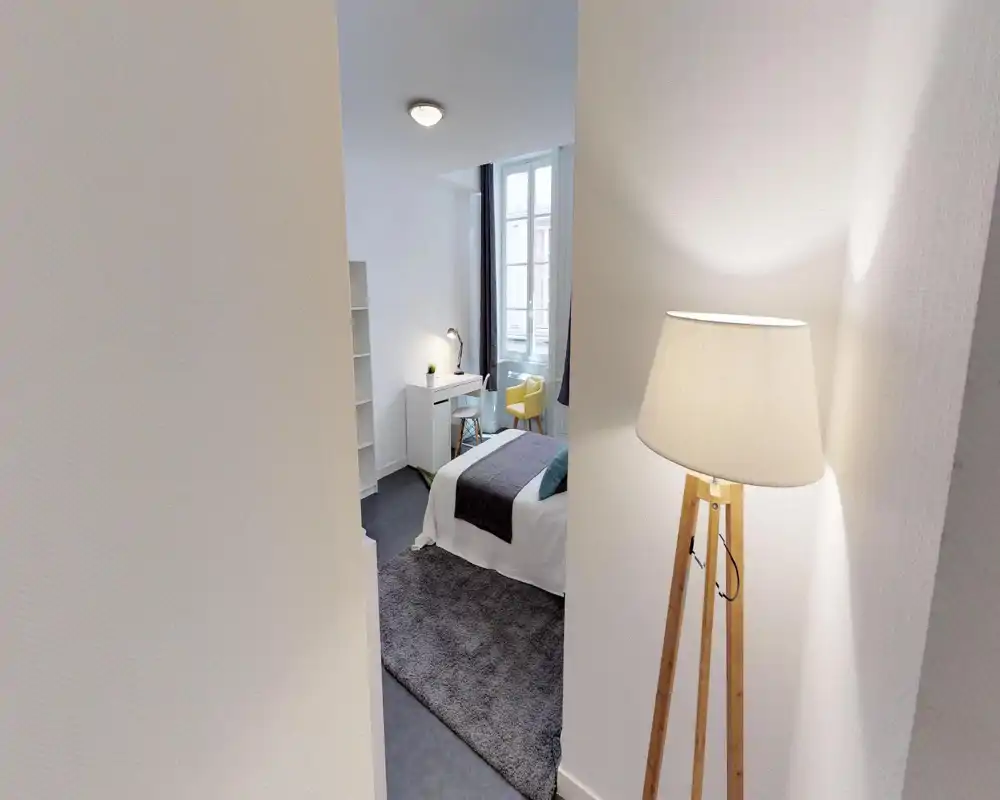 Dormitorio moderno y luminoso con cama bien hecha, alfombra acogedora y rincón de estudio junto a una gran ventana; una lámpara de pie aporta calidez.
