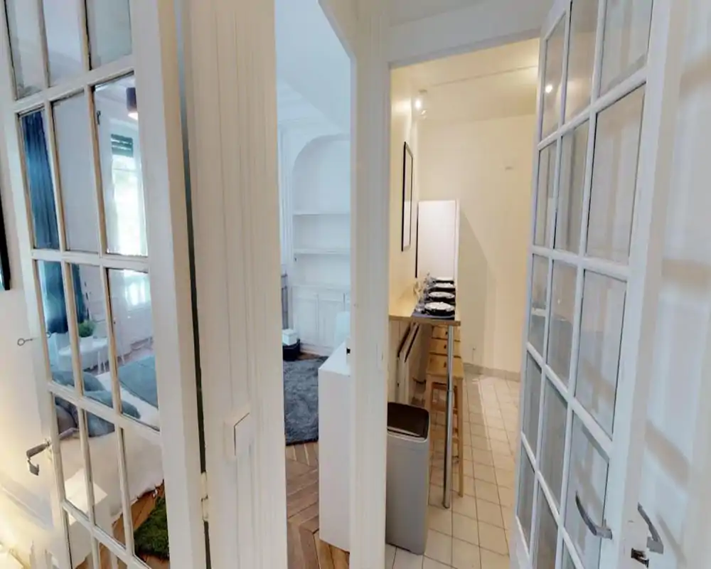 View from a corridor into a bright, furnished living area and a small breakfast bar/kitchenette; white framed glass doors, rug and soft textiles create a cozy atmosphere.