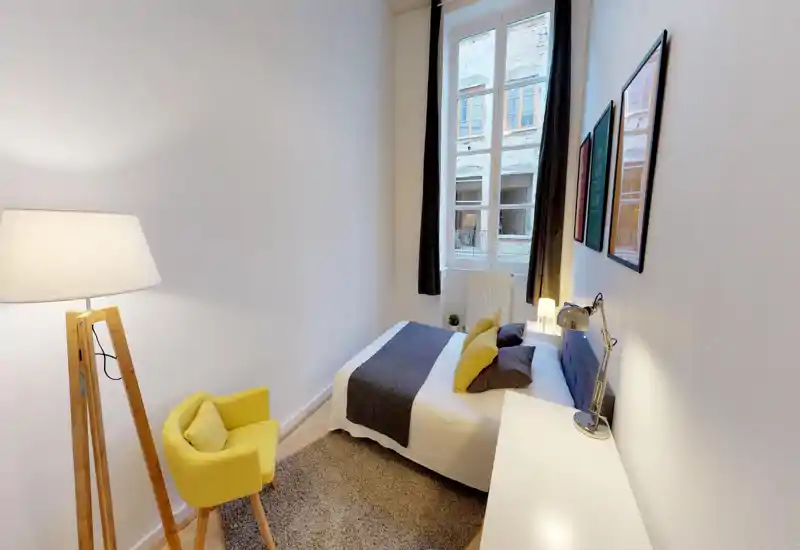 Bright, compact bedroom with a double bed dressed in neutral linens, decorative pillows and a bedside lamp. A large window brings natural light and the yellow accent chair and framed prints add cheerful, modern touches.