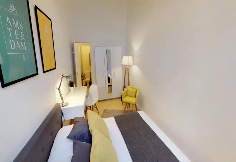 Bright, modern bedroom with a large bed in the foreground, a study desk and chair along the wall, wardrobe with mirror and a cozy yellow armchair with floor lamp.