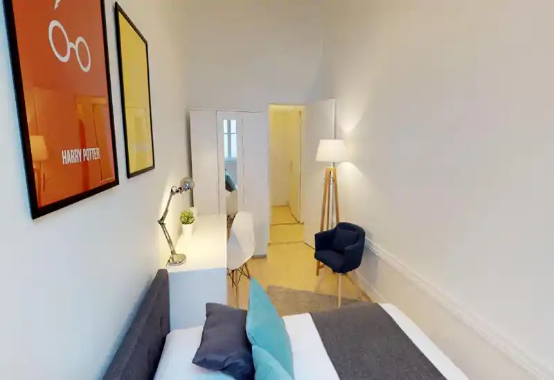 Bright, well-staged bedroom with a double bed, bedside desk and lamp, wardrobe and a cozy reading chair. Clean modern decor with colorful cushions and framed prints.