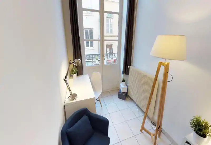 Bright, well-styled small living area with a comfortable armchair, floor lamp, desk and large window letting in natural light — ideal for relaxing or working.