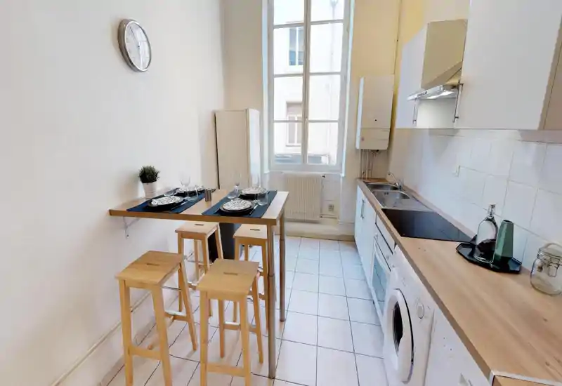 Bright, compact kitchen with a wooden worktop, induction hob, sink and washing machine. A tall window provides natural light and a small high table with stools creates a casual dining area.