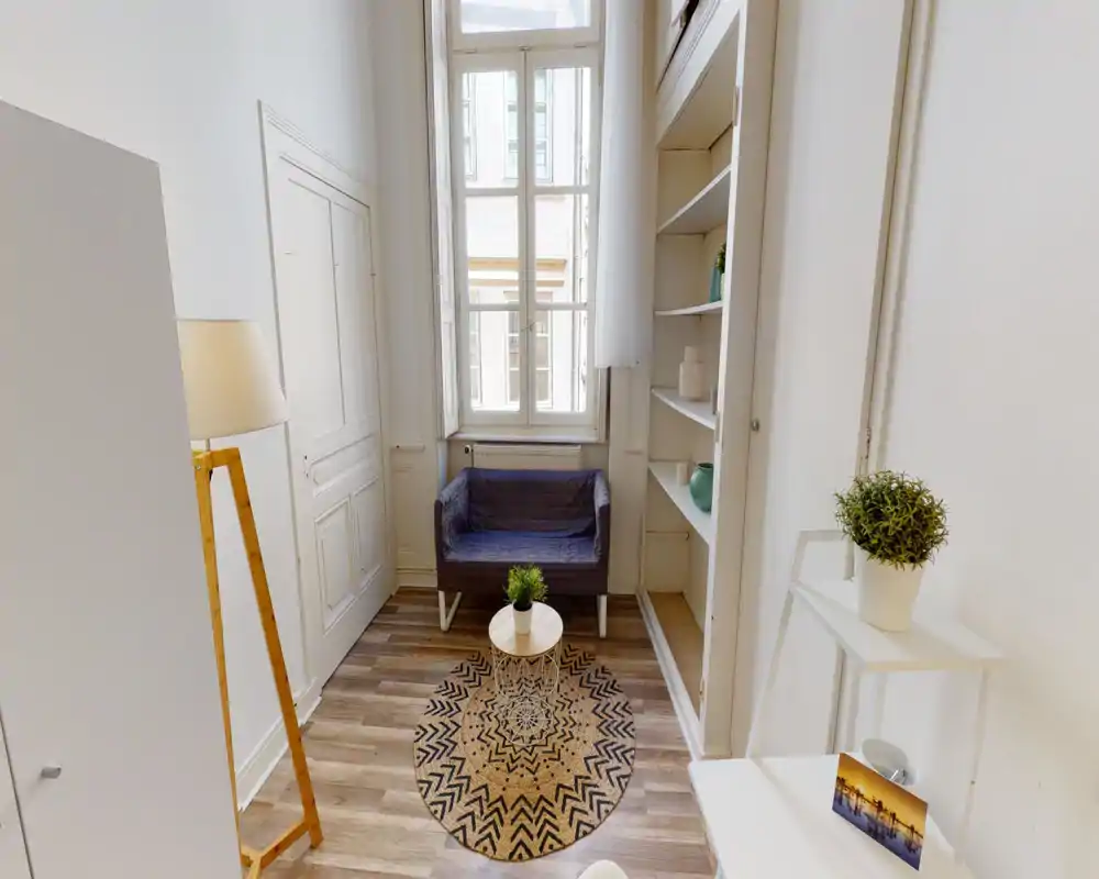 Small, bright living area with a compact two-seater sofa, decorative round rug and natural light from a tall window — cozy, simply styled lounge corner ideal for relaxation.