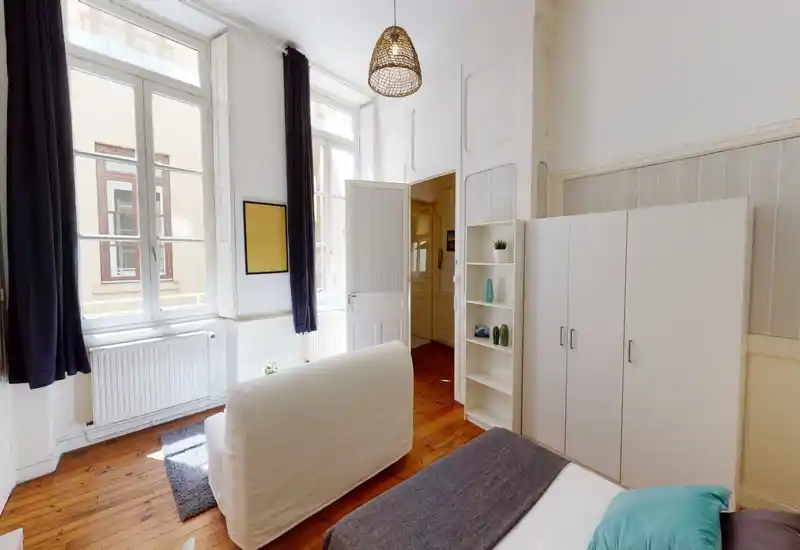 Bright, stylish bedroom with large windows, wooden floors and a comfortable bed. Thoughtful décor and high ceilings create an inviting atmosphere ideal for rental listings.