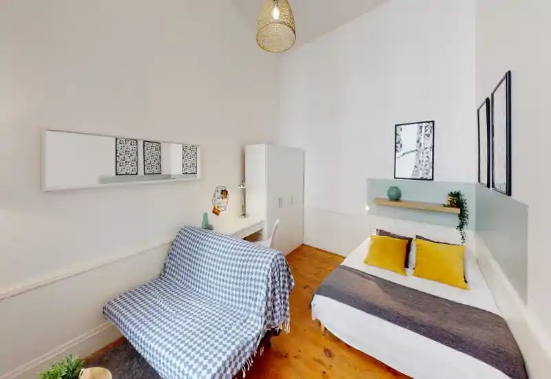 Bright, stylish bedroom with a comfortable double bed dressed with mustard cushions, a small desk and wardrobe, and a cozy seating area with patterned throw — ideal for a single occupant or couple.