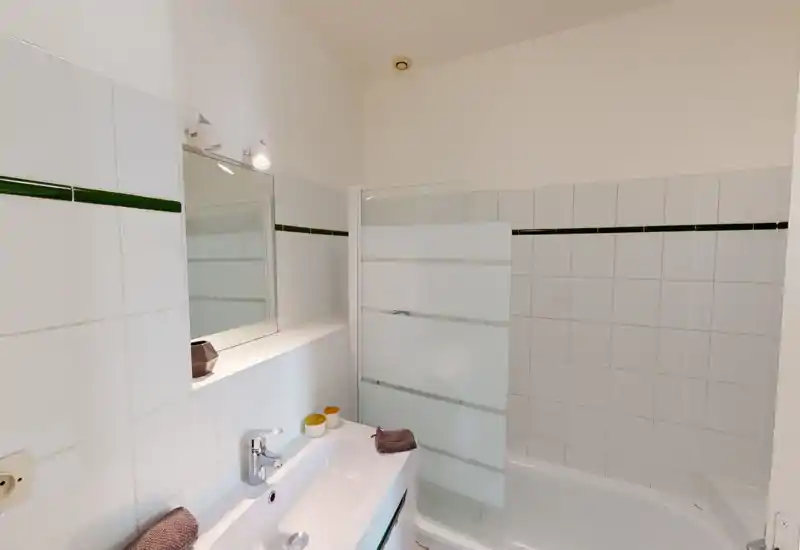 Salle de bains propre et lumineuse avec lavabo blanc, robinet chromé, murs carrelés et baignoire équipée d'un pare-douche en verre dépoli.