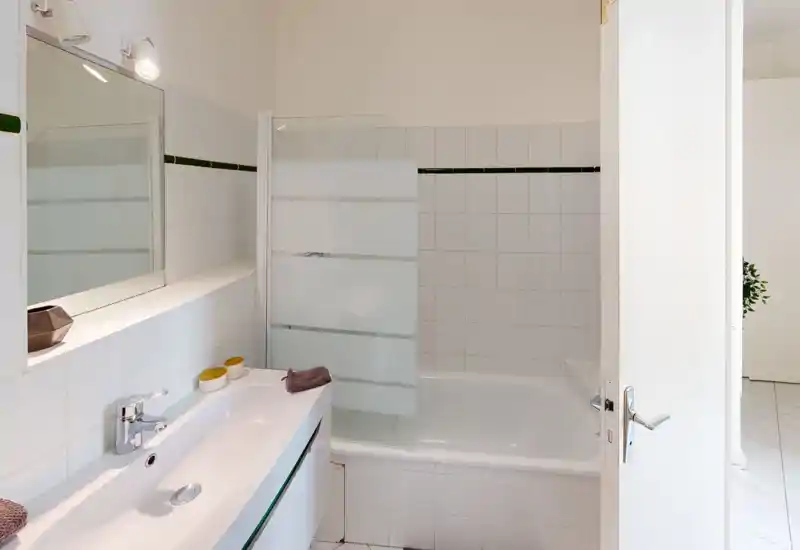 Salle de bains blanche et propre avec lavabo, miroir et baignoire munie d'un pare-douche en verre dépoli. Luminosité et style neutre, idéal pour une annonce locative.