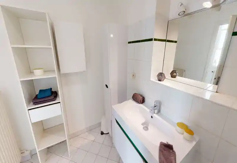 Salle de bains lumineuse et propre avec lavabo moderne, grand miroir et murs carrelés ; étagères avec serviettes pliées pour le rangement.