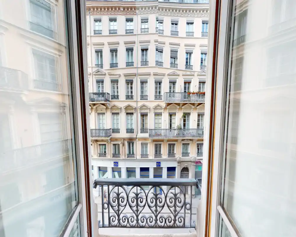 Small balcony view overlooking a classic multi-story building façade; decorative wrought-iron railing and open windows provide a charming urban outlook.