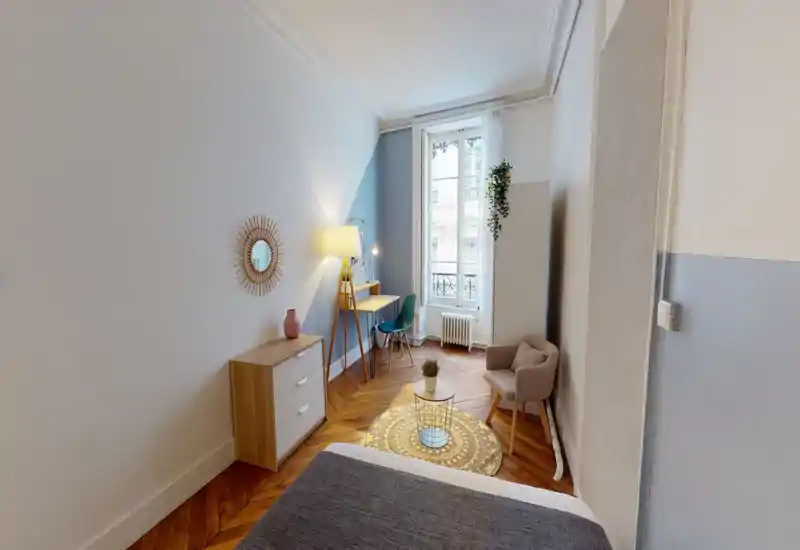 Camera da letto luminosa e di stile con letto ben visibile in primo piano, parquet in legno, una poltrona accogliente e una scrivania vicino a una grande finestra che lascia entrare luce naturale.