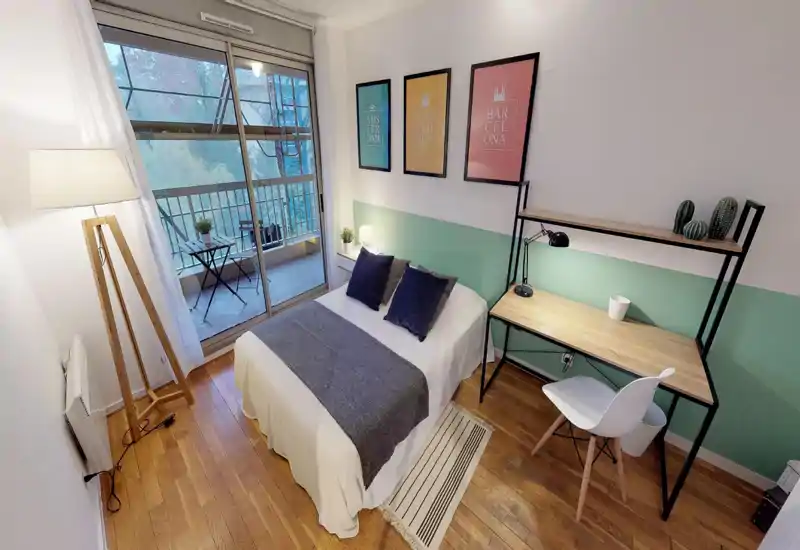 Bright, well-styled bedroom with a double bed, bedside table and a desk. Large sliding doors open to a small balcony, flooding the room with natural light.