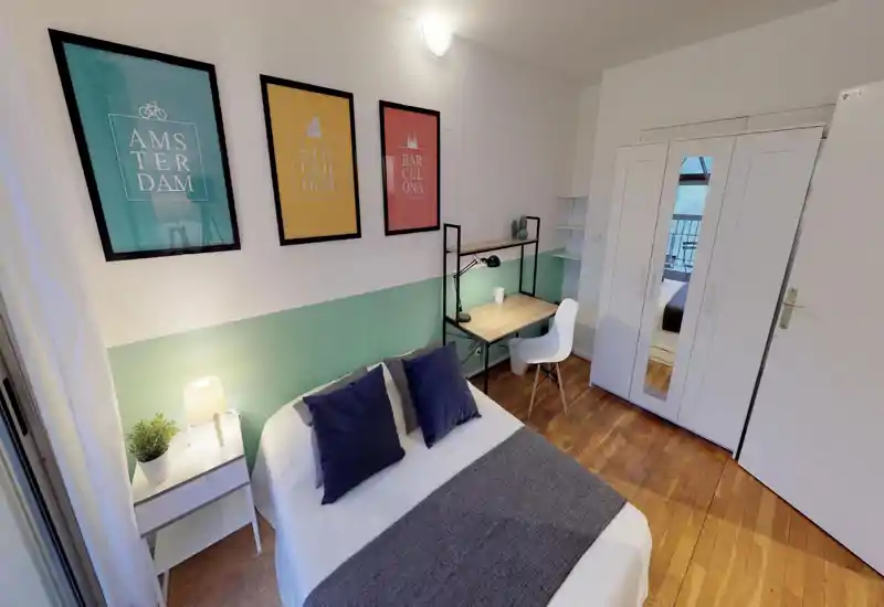 Bright, neatly styled bedroom with a double bed, bedside table with lamp, a desk and wardrobe; calm pastel wall accent and framed prints give a cozy, modern feel.