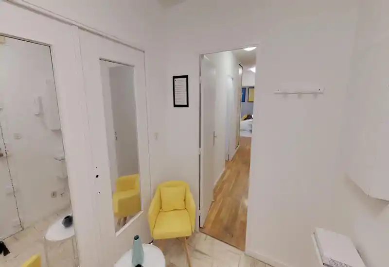 Small, well-lit entrance/foyer with a bright yellow accent chair, wall mirror and a side table; wooden corridor leads deeper into the apartment.