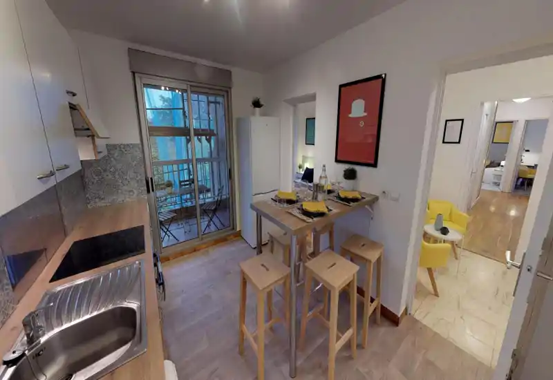 Compact modern kitchen with a wooden countertop, stainless sink, induction hob and a small high dining table set for four; sliding doors open to a balcony with outdoor seating.