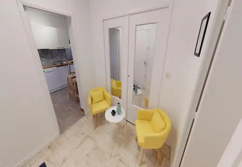 Small, bright entrance area with two yellow accent chairs, a round white side table and mirrored closet doors; visible doorway leads to the kitchen. Clean, modern styling and warm accents make the entry welcoming.