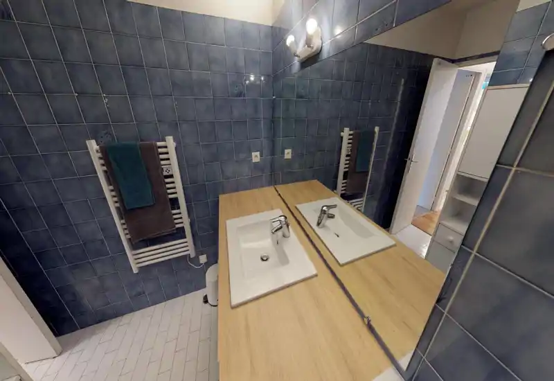 Modern bathroom with blue tiled walls, a large mirror and a double sink on a wooden countertop; heated towel rail with folded towels and tiled white floor.