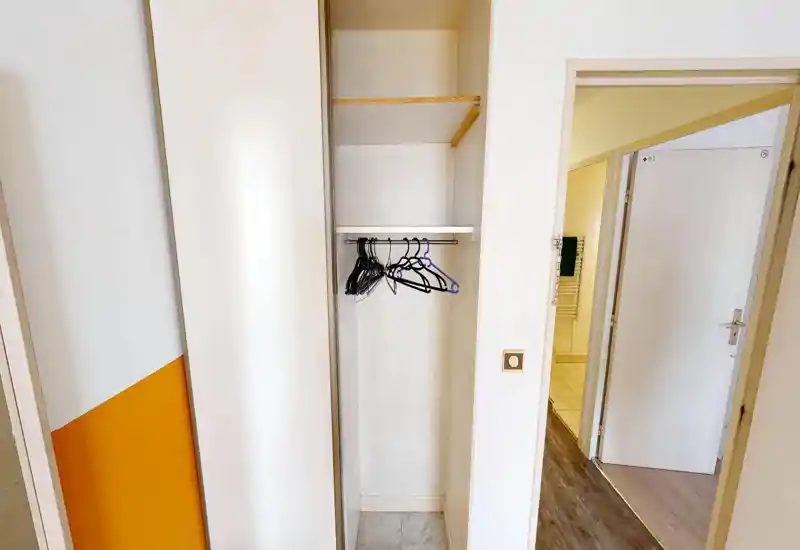 Petit espace d'entrée/placard avec penderie intégrée et quelques cintres ; murs blancs neutres et porte donnant sur d'autres pièces.