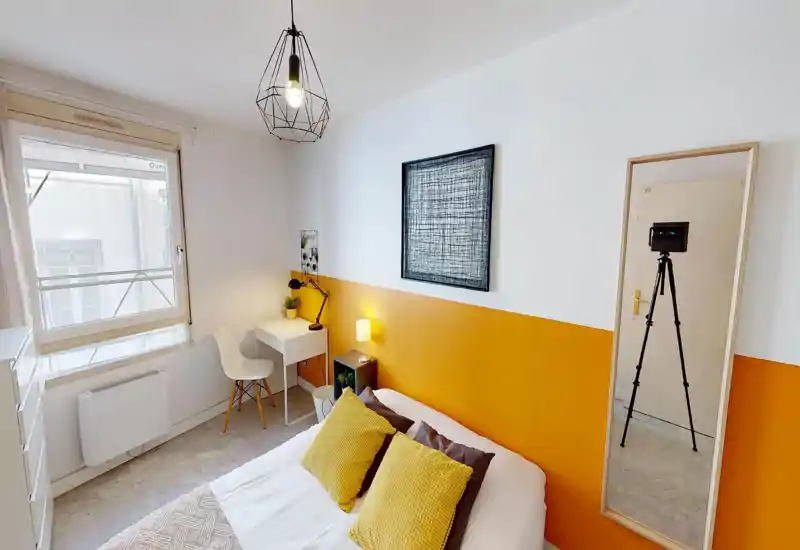 Chambre moderne et lumineuse avec lit double habillé de coussins jaunes, petit bureau près de la fenêtre, lampe de chevet et tableau décoratif. Espace bien éclairé et soigné, parfait pour une annonce.