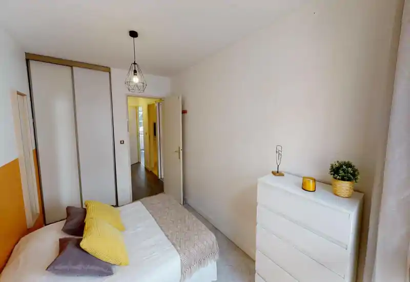 Chambre lumineuse et accueillante avec un lit double bien fait, coussins jaunes, placard intégré et commode blanche — idéale pour un séjour confortable.