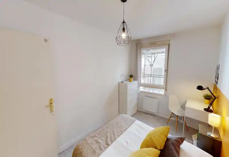 Chambre lumineuse et soignée avec lit bien visible, textiles doux et accents jaunes chaleureux. Un bureau et une chaise près de la fenêtre; la pièce est bien éclairée par la lumière naturelle et des lampes.
