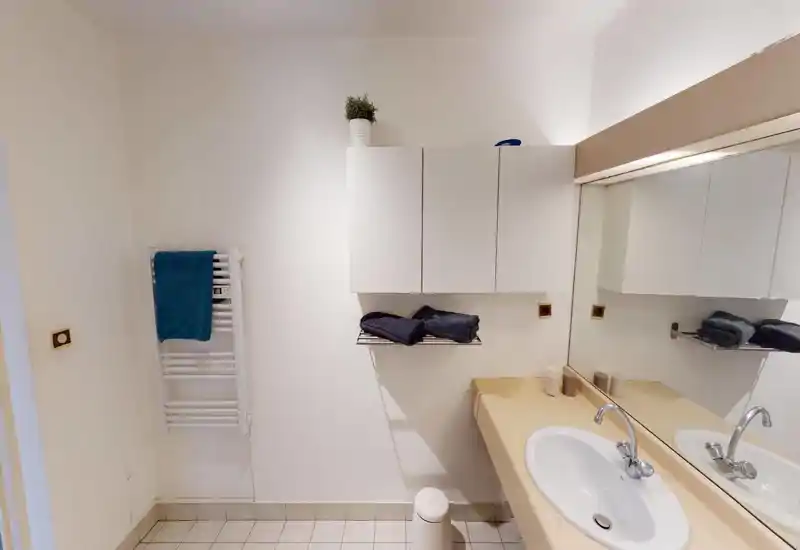 Salle de bains propre et minimaliste avec lavabo encastré, grand miroir, placards muraux et porte-serviettes. Tons neutres et serviettes pliées pour un espace soigné et fonctionnel.