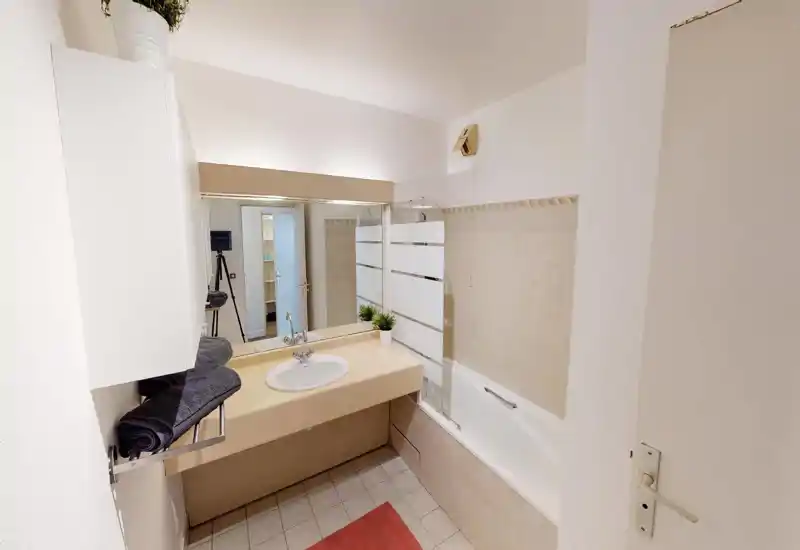 Salle de bains propre et lumineuse avec baignoire et lavabo intégré, carreaux neutres et grand miroir qui agrandit visuellement l'espace.